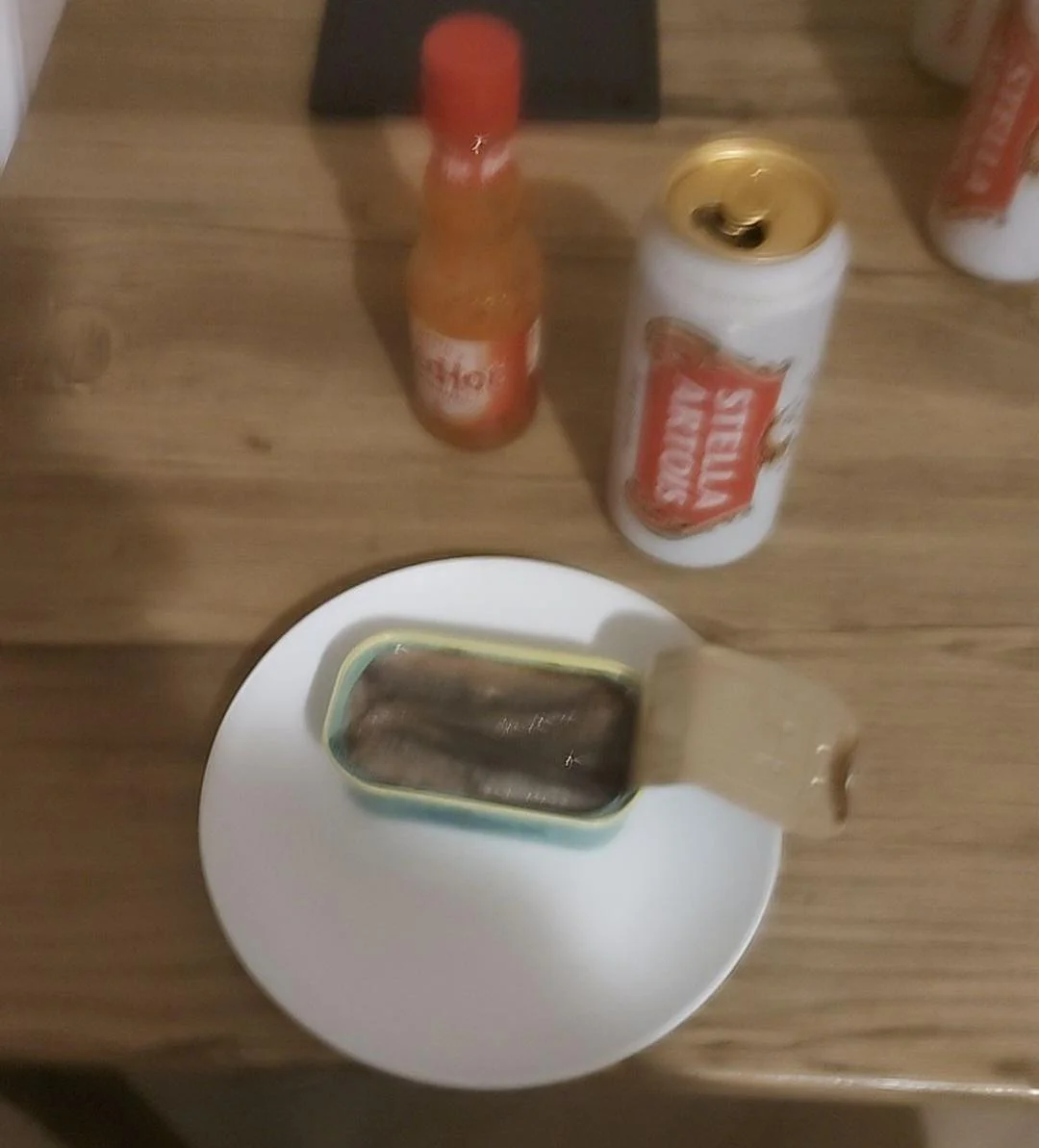 A blurry photograph of an opened tin of sardines on a clean white plate, flanked by a bottle of Frank's Red Hot and an opened can of Stella Artois