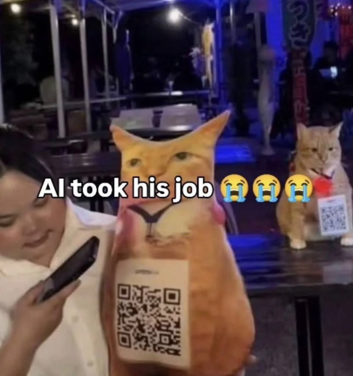 A stuffed cat with a QR code printed on him.  In the background is a real cat with a qr code on a lanyard around his neck.  "AI took his job!"